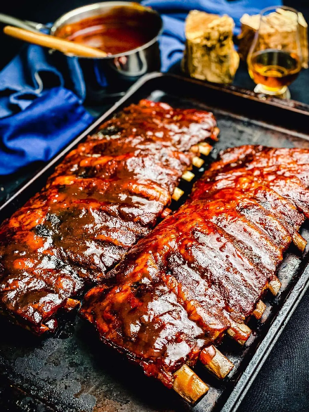 Two racks of glazed, caramelized competition BBQ ribs on a dark sheet pan with a pot of sauce